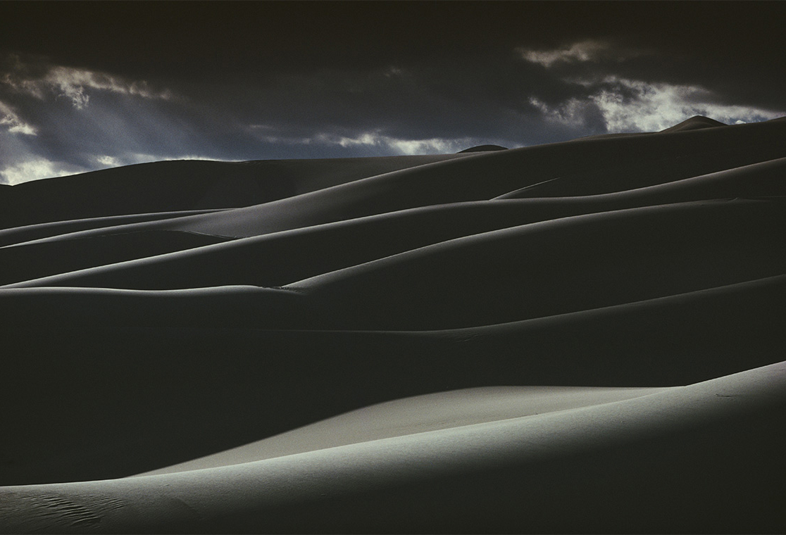 Dark sand dunes lit only along their crests, falling in repeating curved ridges across the frame, with a narrow strip of cloudy blue sky at the top.