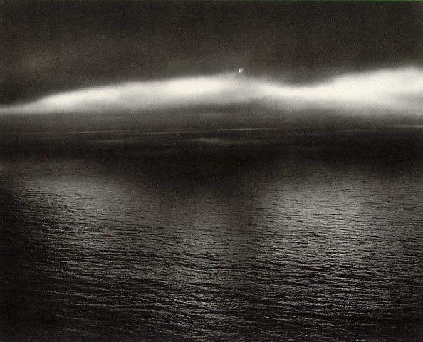 Dark moody seascape with a soft luminous cloud band hovering over a textured dark sea, a small bright point of light at the cloud's crest.