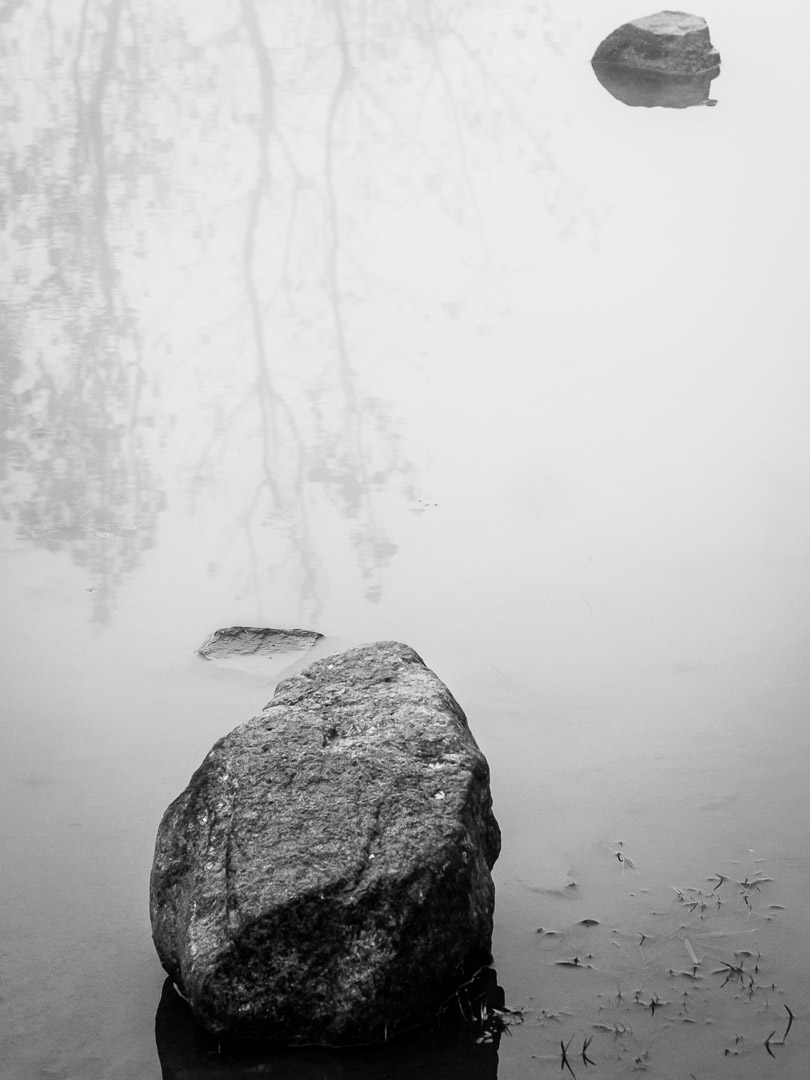A single dark boulder rises from misty still water, with faint reflected tree branches in the upper left.
